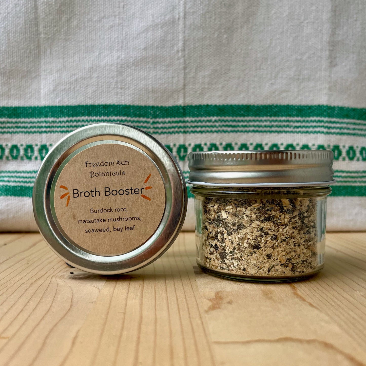 Two jars labeled 'Broth Booster' on a wooden surface with a striped fabric background.