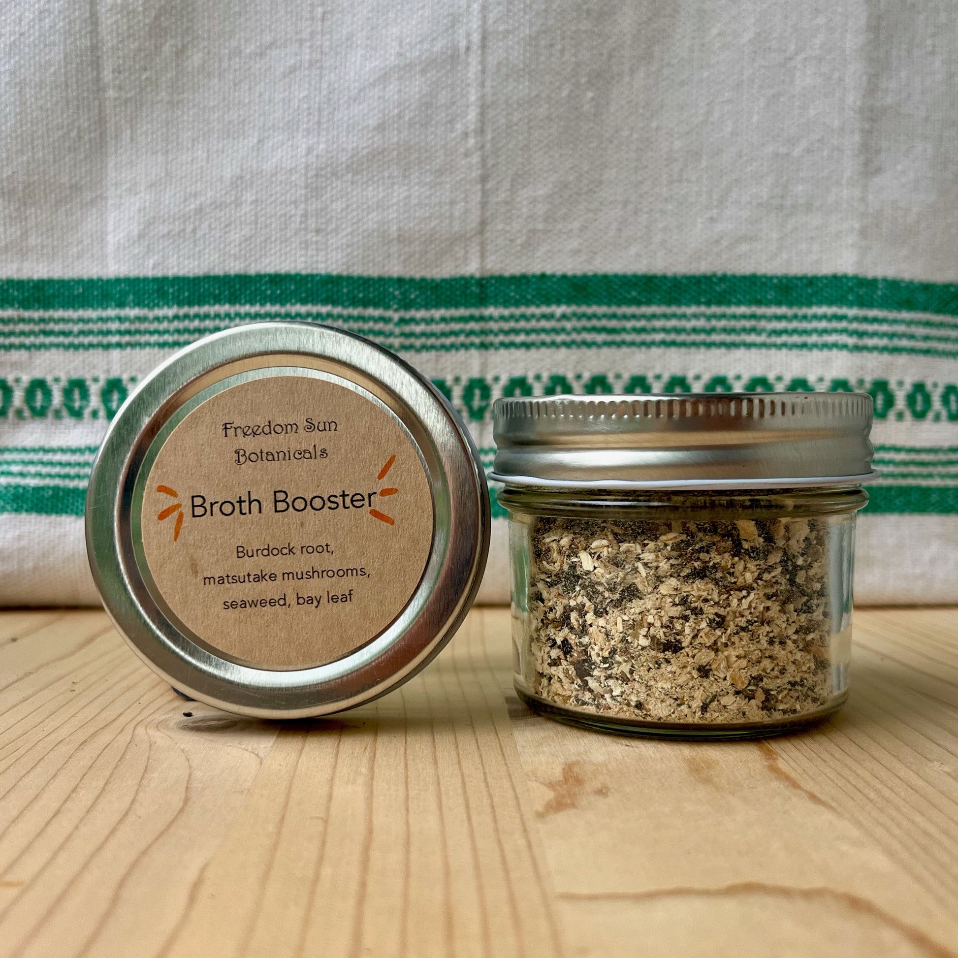 Two jars labeled 'Broth Booster' on a wooden surface with a striped fabric background.