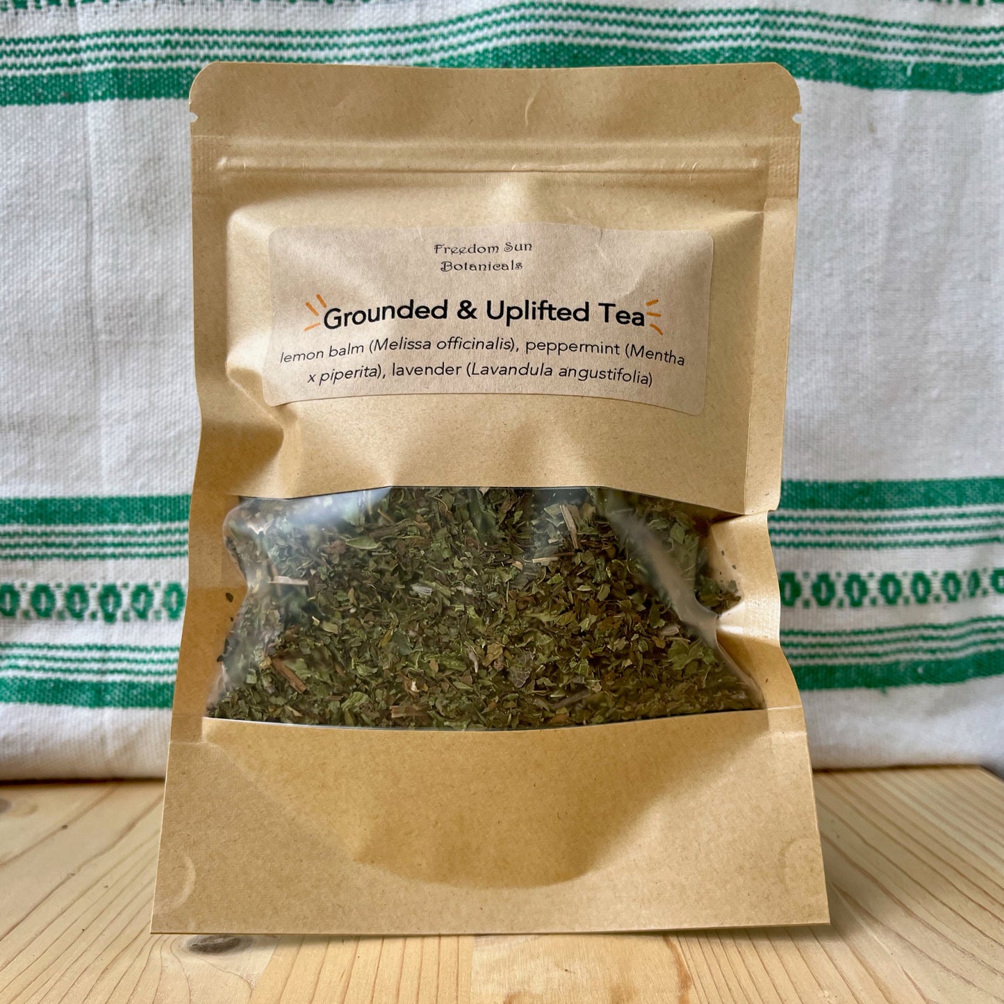 Tea package labeled 'Grounded & Uplifted Tea' on a wooden surface with a green and white striped cloth in the background.