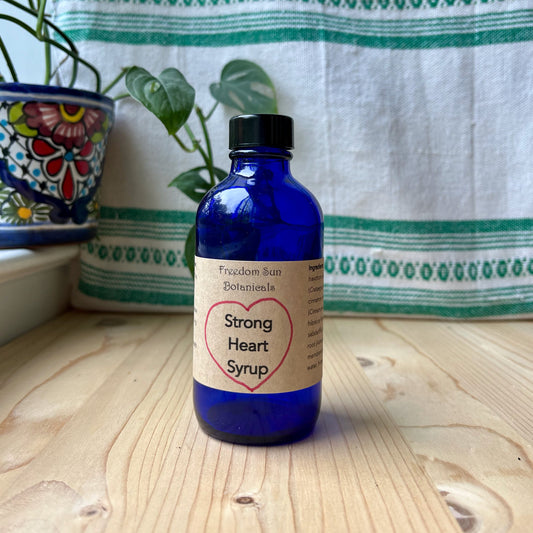 Blue bottle of 'Strong Heart Syrup' on a wooden surface with a plant and fabric in the background.