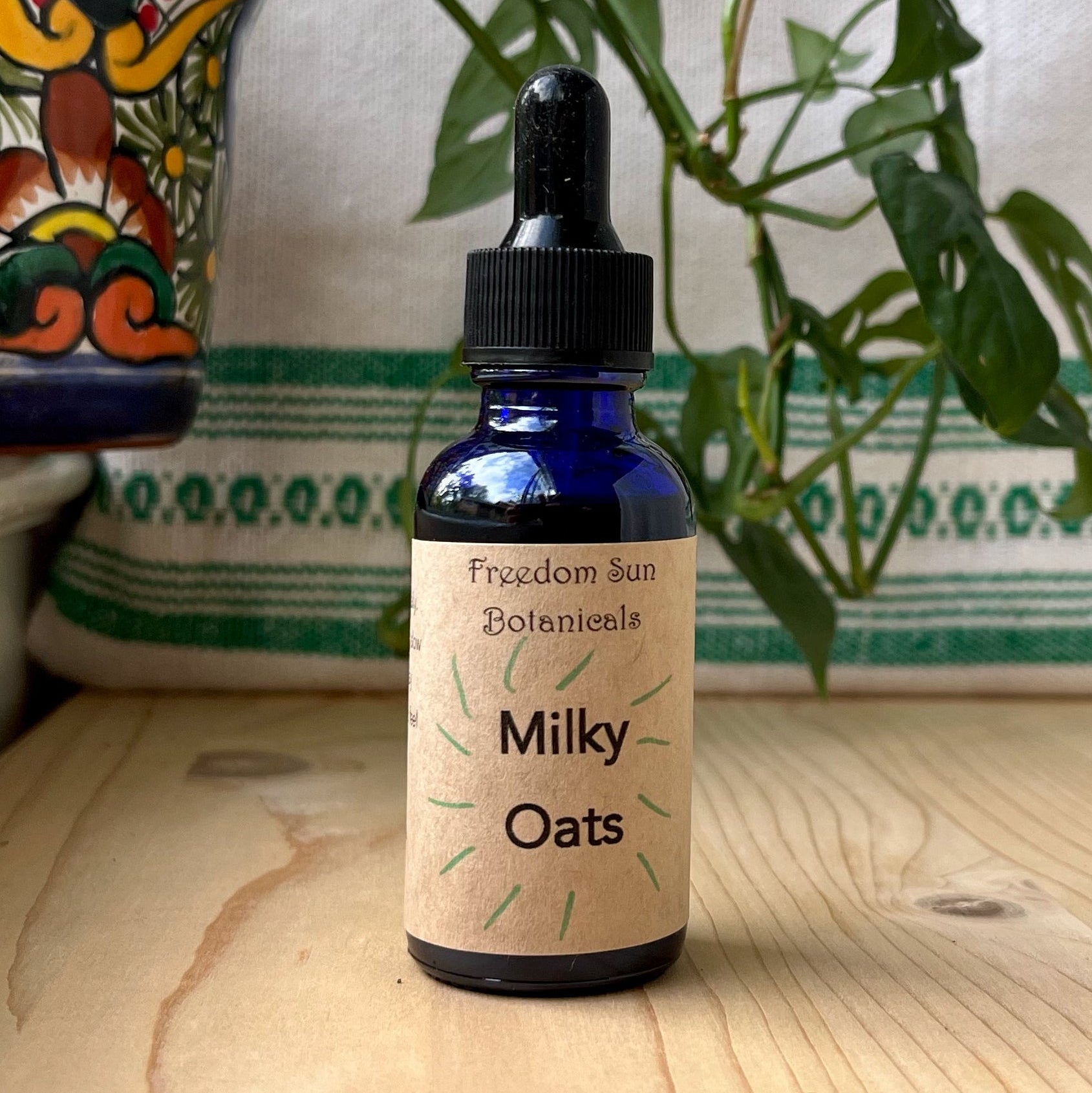 Bottle of 'Milky Oats' with a dropper on a wooden surface with a plant and decorative item in the background.
